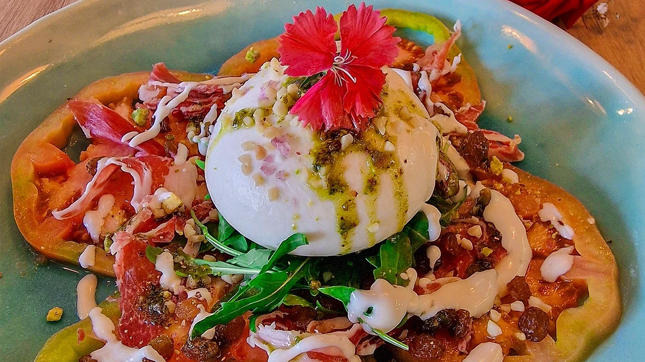 Doña Burrata Carpaccio de tomate rosa, con burrata fresca, pesto de pistacho, rúcula, jamón ibérico, pasas, almendras, AOVE y vinagreta de Yogurt griego del Restaurante Azzurro